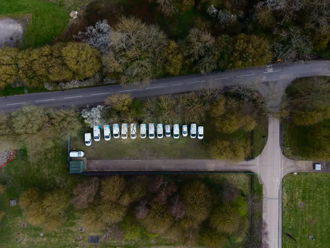 Vista aérea de um pátio de garagem com quinze veículos de frota estacionados em fila junto à estrada.